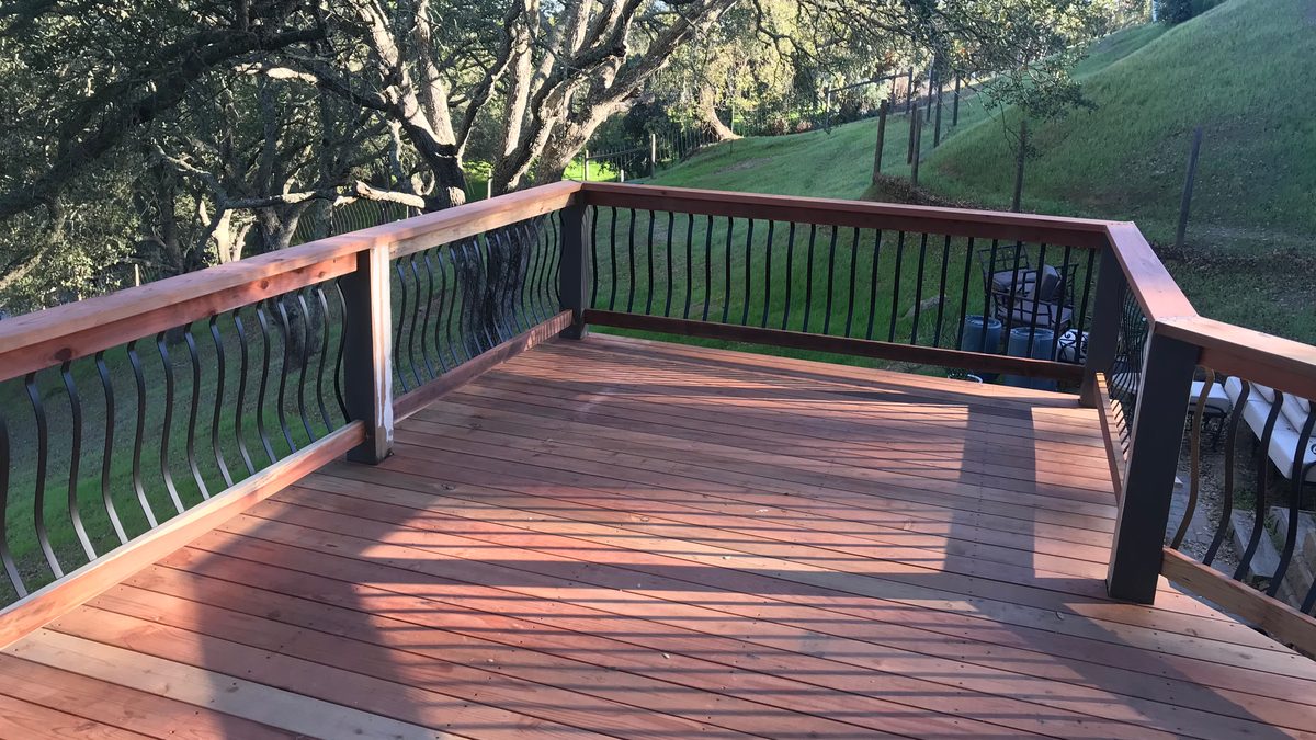 A stained wood deck with a dark metal baluster railing, overlooking a green hillside.