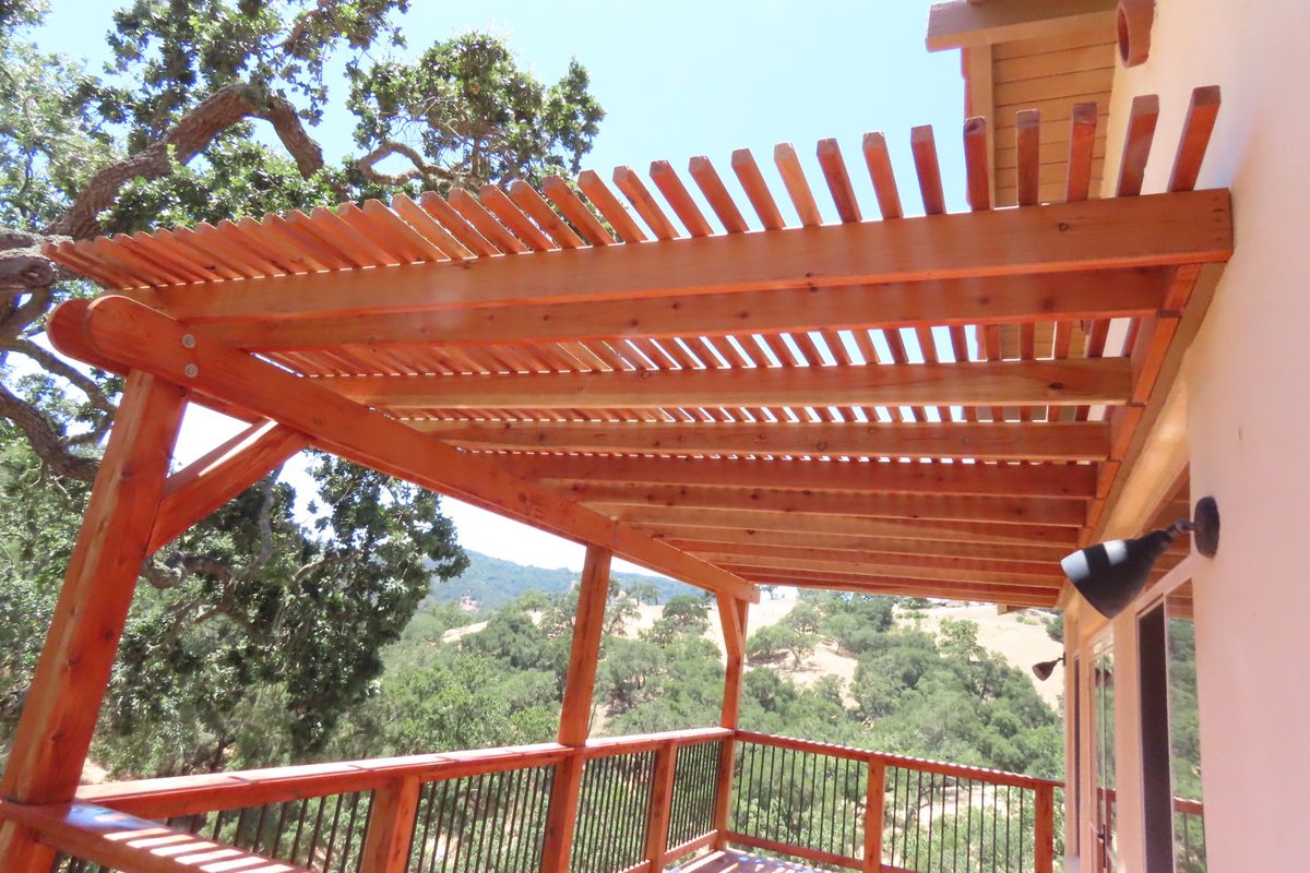 A stained wood pergola with closely spaced rafters over a deck with a dark metal railing, oak-covered hills beyond.