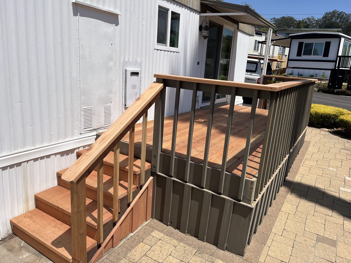 New wooden steps and a small landing with a railing, against a white-sided home on a paver patio.